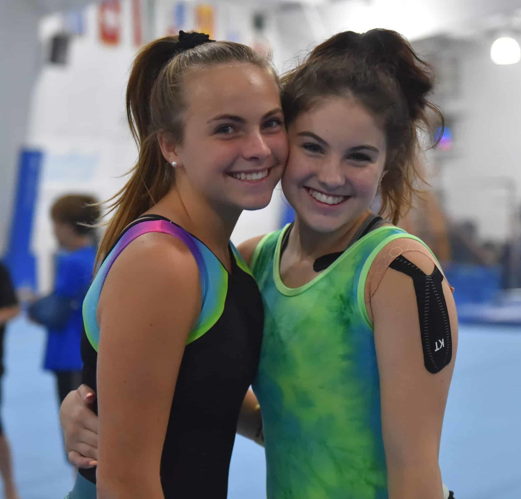 Two gymnasts pose for a photo. They're wearing gymnastics attire and one has athletic tape on her shoulder.
