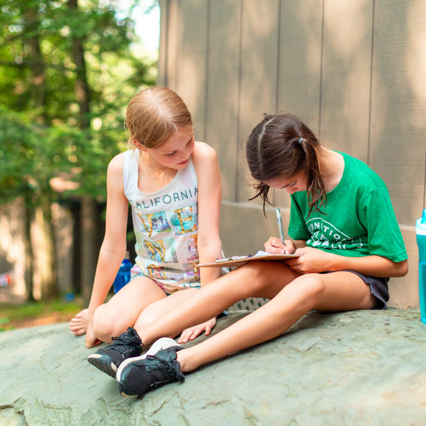 two campers sitting down writing on a clipboard